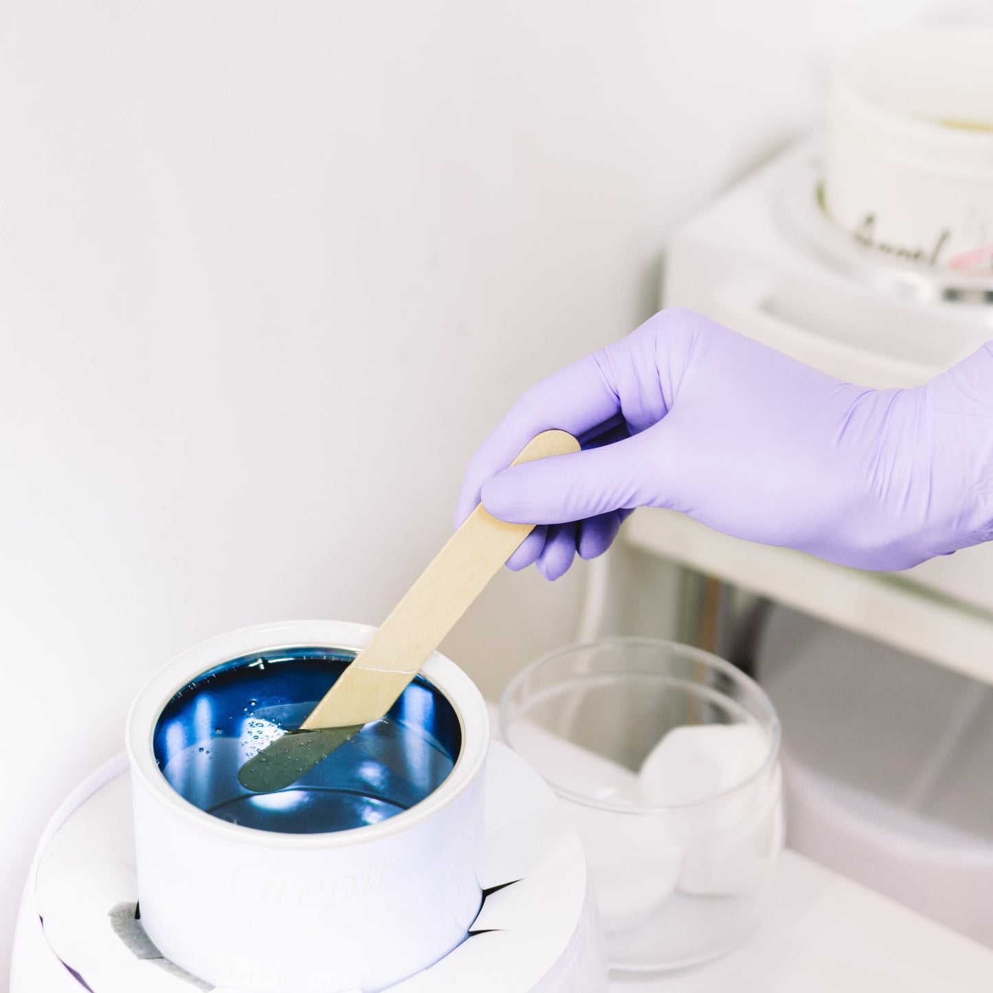 A person wearing a purple glove is holding a waxing tool over a container of blue wax, about to apply the wax on a client's leg.