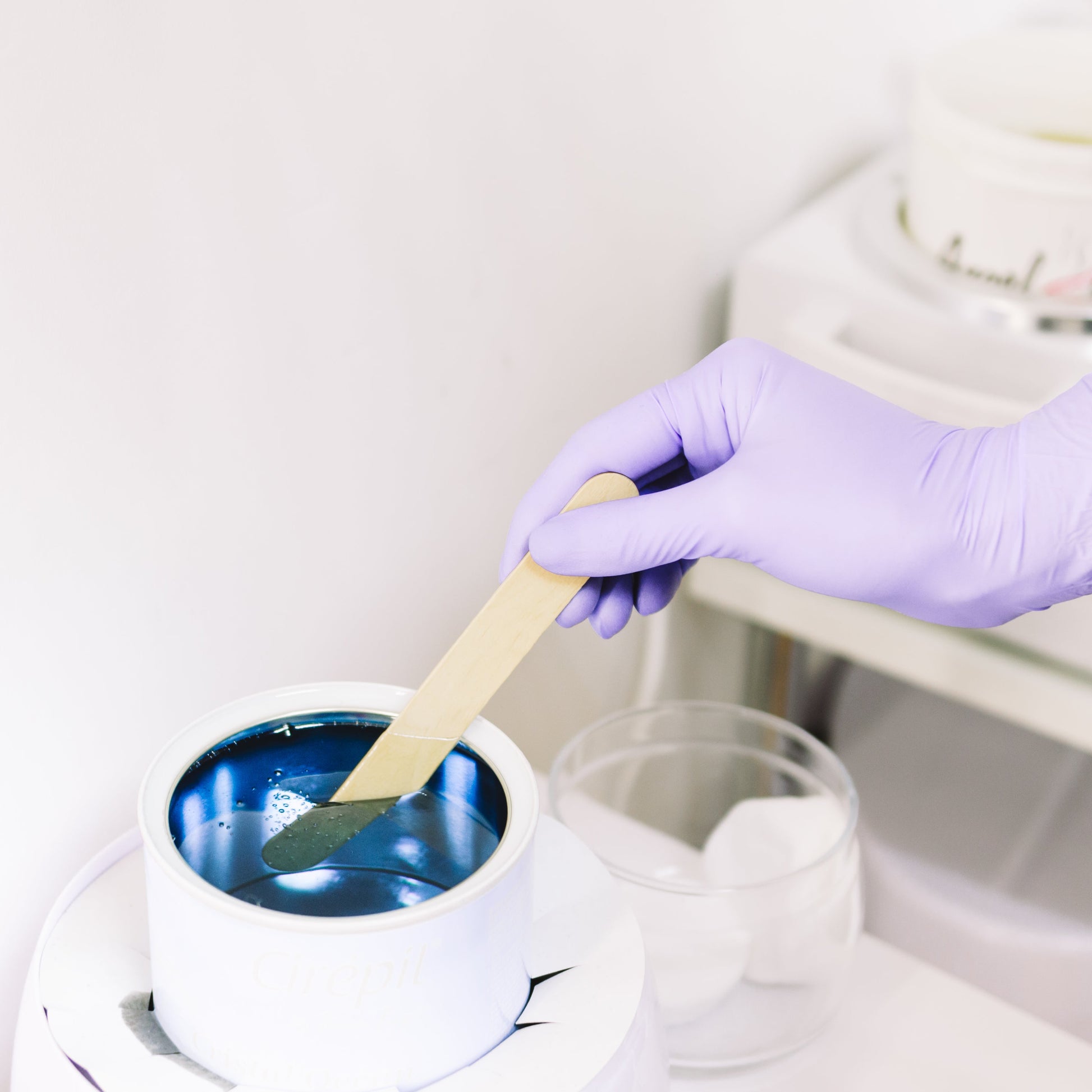 A person wearing a purple glove is holding a waxing tool over a container of blue wax, about to apply the wax on a client's leg.