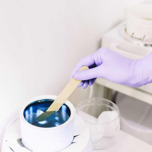 A person wearing a purple glove is holding a waxing tool over a container of blue wax, about to apply the wax on a client's leg.